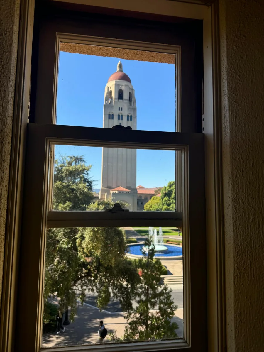 Hoover Tower against a clear blue sky, seen through an auditorium window at Stanford, with campus trees and a fountain below.